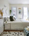Bright bedroom with a white bed, green pillows, desk, and wall art, featuring large windows and a modern floor lamp.