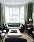 Elegant living room with green walls, white armchairs, a backgammon table, and a bay window with shutters and curtains.