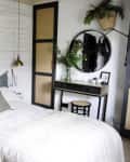 Cozy bedroom with white shiplap walls, green plants, a round mirror, and a black vanity table with a stool.
