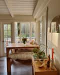 Sunlit dining area with wooden table, fur-covered bench, vase of flowers, and a red candle on a side table.