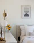 Cozy bedroom with a white bed, abstract art, yellow flowers, and a modern lamp on a dresser.