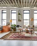 Loft living room with large windows, eclectic rugs, wooden furniture, plants, and modern decor.