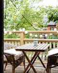 Wooden balcony with wicker chairs, a small table, string lights, and lush green trees in the background.