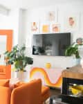 Bright living room with orange sofa, large mirror, plants, wall art, and a mounted TV.
