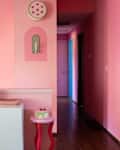 Pink hallway with framed abstract art, a white cabinet with books, and a small table holding a decorative cake.