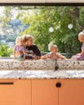 Family enjoying a sunny day by a large kitchen window with terrazzo countertop and orange cabinets, overlooking a lush garden.