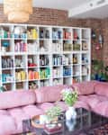 Exposed brick living room with pink tufted sofa, glass coffee table, and a large bookshelf organized by color.