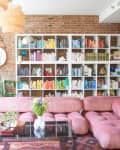 Pink tufted sofa in a living room with a brick wall, colorful bookshelves, plants, and woven pendant lights.