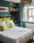 Cozy bedroom with teal walls, white bed, yellow checkered pillows, bookshelf, wicker chair, and large window with curtains.