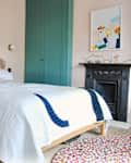 Bedroom with a rattan headboard, white quilt, teal wardrobe, abstract art, and a colorful round rug.