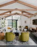 Contemporary living room corner with large white couch, green side chairs, all-black fireplace, glass and black coffee table, silver arched floor lamp, large palm plant, in a room with white ceilings, natural wood rafters, and large windows overlooking a lake.