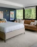 Modern bedroom with large windows, brown leather sofa, upholstered bedframe, and dark blue accent wall behind the bed.