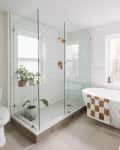 Philadelphia residence with white walls, lots of wood details: bathroom with oval freestanding tub, brown and white checkered bath mat, glass shower with white tiles, rose gold hardware, plants. Window over tub and in shower