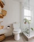 Philadelphia residence with white walls, lots of wood details: bathroom with oval freestanding tub, brown and white checkered bath mat, glass shower with white tiles, rose gold hardware, plants. Window over tub and in shower