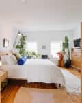 View into bedroom with bed with wood headboard, white linens, blue and green pillows. Lots of plants