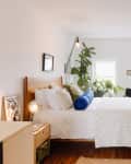 View into bedroom with bed with wood headboard, white linens, blue and green pillows. Lots of plants