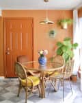 Dining room with bright orange walls, large windows with floor to ceiling white curtains, lots of potted plants, cane round dining table with glass top, cane and rattan dining chairs, blue and white vase of flowers, marble or marble look checkered floor