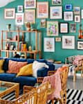 Living room with teal green gallery wall, high ceilings with wood beam, white round pendant light, black and white striped rug, blue velvet sofa with warm colored-accent pillows, cane chairs and coffee table (with glass top), wood shelves with books, art objects