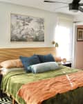 bedroom with pale neutral walls, bed with orange and green linens, bold black and white geometric patterned rug, sliding door in muted blue/green, black ceiling fan, vintage print of plants, bird over bed, slatted wood headboard, deep purple bedside table