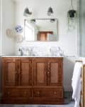 Two sconces above gold frame mirror hung above wooden vanity with marble top in newly renovated bathroom with a grey tiled bathroom.