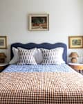 Blue upholstered scalloped headboard in bedroom with neatly made bed with brown and white gingham quilt. Small portraits hung around bed with   straw pendant hung above bed.