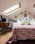 Art prints mounted behind neatly made bed with pink floral bedding in bedroom and writing desk sitting on wall underneath one of two skylights. Small built in storage unit on other side of bed.