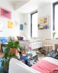 Bed in corner of studio apartment with colorful artwork lining the wall. Small desk with cantilever chair underneath sits beside table.