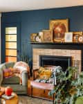 Blue painted wall in living room with ombre glass yellow to orange glass entryway door and various armchairs topped with vintage blankets. Vintage floral paintings and prints displayed on fireplace mantel.