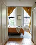 Neatly made orange bedding in light filled bedroom with yellow drapes over large windows.