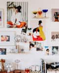 Gallery wall with colorful art prints, photos, and glassware on shelves above a table with books and a coffee maker.