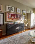 Living room with sage/olive colored walls, gallery wall, black credenza, 2 peach armchairs with throw pillows, gray patterned rug, gray wardrobe/cabinet, patterned floor to ceiling curtains, sofa with patterned throw pillows, glass and gold oval coffee table