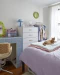 A bedroom with one pink feature wall. A blue tiled desk is next to a white dresser. On the bed is a purple duvet with an orange cat laying down on it.