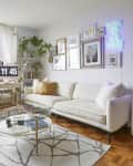 Bright living room with white sofa, round chair, glass coffee table, wall art, neon sign, and plants by a large window.