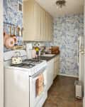 Narrow kitchen with blue floral wallpaper, white cabinets, gas stove, and hanging copper pans.