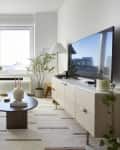 A narrow living room with a white couch, light wood TV stand, and a large window