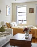 Cozy studio apartment with a white sofa, green pillows, wall art, a bed with yellow bedding, and a desk by the window.