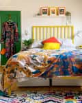 bedroom, yellow slated headboard, angled ceiling, green doors, plants, colorful comforter, multi-colored rug, wood floors, sunlight
