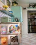 white and pink large geometric tile floor, floating book display shelves, mint green refrigerator, mint green toaster, doorway, floating shelves, multi colored books, mint stand mixer, pink and white geometric tile floor, green and pink color blocking, natural wood cabinets, white hood vent, white oven, teal art deco backsplash, floating shelf, glass panel door, blue countertops, pink tea kettle, green and pink rainbow curve wall sconce with white bulb