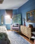 One wall of bedroom. Wall is blue with wainscoting, credenza with cool design, painting of woman with gold frame, velvet lilac armchair, blue screen with blossoming trees, blue shag rug on wood floor, bed has pink and pale blue bedding, faux fur throw. Large windows in back with green curtains