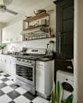 kitchen with white cabinets, black and white checkered floor, white tile backsplash, blue walls with white trim. Shelves over stove area with spices, pans. Plant leaves in white pitcher on counter. To the left is the dining area with small round marble bistro table with clear lucite chairs. Black and white checkered floor, blue walls and radiator with white trim. Pots and pans hanging above dining table. 2 windows letting in light