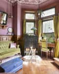 living room with pink walls, pink and white decorative ceiling, tall yellow/green curtains, green velvet antique sofa with shiny pink velvet pillow and throw, wood floors, 3 large windows, antique desk with pale green chair