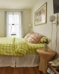 Bedroom with bed with chartreuse and white gingham duvet with floral and plaid pillowcases, straw round bedside table with case of yellow flowers, wood shoe rack with wall mount TV over it, windows with sheer white curtains, wood floor, white dresser with arched mirror across from bed