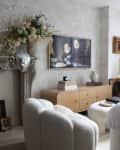 Television with renaissance image above blonde wood media credenza in light filled living room with neutral colored furniture. Fireplace mantel placed on wall with large urn floral arrangement.