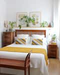 bedroom with white walls, bed with mustard, gray and white bedding with wood headboard, wood side tables, shelf over bed with plants, art, wood slat bench at foot of bed, wood floors