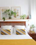 bedroom with white walls, bed with mustard, gray and white bedding with wood headboard, wood side tables, shelf over bed with plants, art