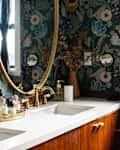 Floral wallpaper in bathroom with wooden vanity topped with white stone surface. Vanity surface in lined with perfume collection.