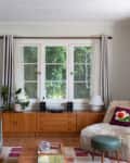 living room with furry chair, with flower pillow potted zz plant, mounted TV, small blue ottoman/stool, large wood credenza, patchwork style rug, wood coffee table, gray velvet sofa, large windows
