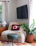 corner of living room with furry chair, with flower pillow potted zz plant, mounted TV, small blue ottoman/stool, large wood credenza, patchwork style rug