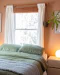 bedroom with peach or pale terracotta walls, bed with pale green bedding, framed art and guitar on wall, arched full length mirror, wood dresser with jewelry, other decor