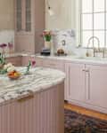 Mauve colored kitchen cabinets in renovated kitchen with white and grey marbled countertop and brass hardware. Capiz shell light fixture above island.